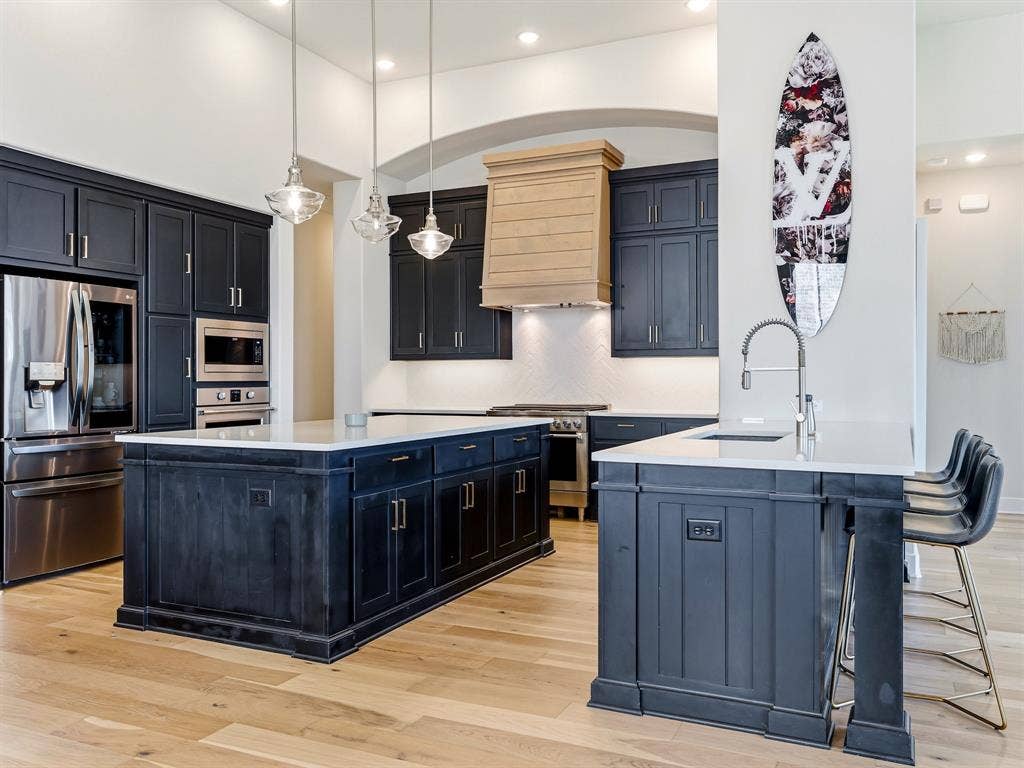 Kitchen featuring charcoal-toned cabinetry, light-toned wood-finish flooring, dual islands with quartz countertops, a custom wood-paneled range hood, and pendant lighting