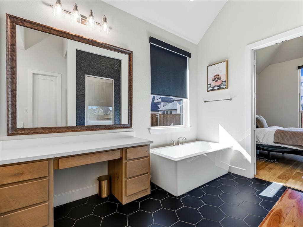 Bathroom featuring black hexagonal floor tile, freestanding soaking tub with chrome fixtures, wood-finish cabinetry, white countertop, and oversized framed mirror