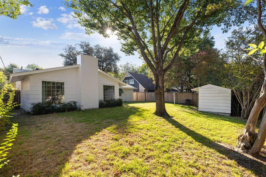 3209 Sheffield Street, Irving, TX 75062 Backyard featuring white horizontal siding, a central masonry chimney, mature trees, a detached storage structure, and wood privacy fencing