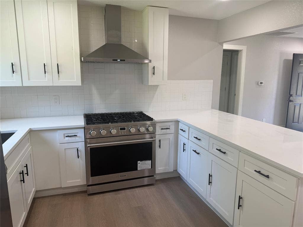 1123 Hansboro Avenue, Dallas, TX 75224 Kitchen featuring white shaker-style cabinetry with matte black hardware, stone-finish countertops, herringbone-pattern tile backsplash, stainless steel gas range, and stainless steel range hood
