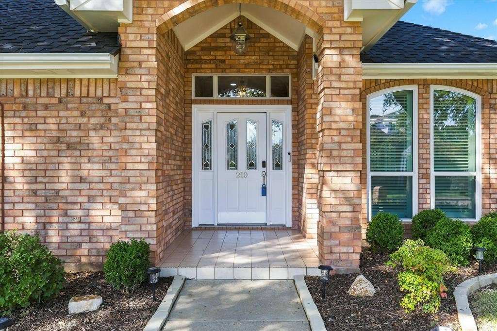 210 Santa Fe Trail, Waxahachie, TX 75165 Brick exterior featuring a covered entryway with an arched portico, a white entry door with decorative glass inserts, an arched transom window, and a tiled porch surface