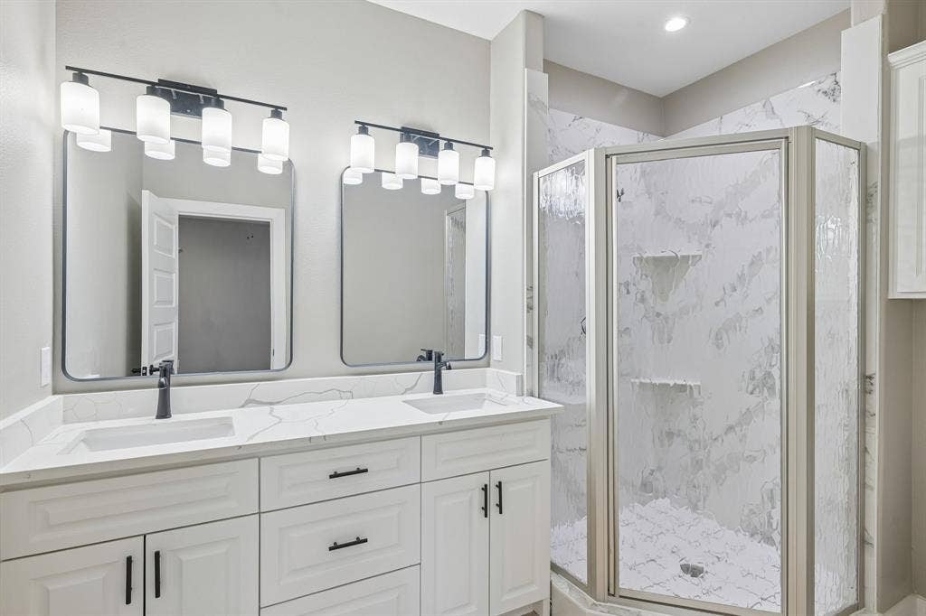 1221 S Rockwall Avenue, Terrell, TX 75160 Dual-sink vanity with a solid-surface countertop and matte black fixtures, paired with two framed mirrors, multi-bulb wall lighting, and a walk-in shower featuring patterned tile surround and glass enclosure