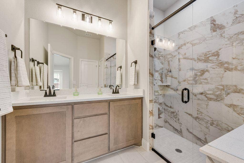 208 Shady Bank Way, Nevada, TX 75173 Double vanity featuring light-toned cabinetry, quartz countertops, dual under-mount sinks, matte black fixtures, and a walk-in shower with floor-to-ceiling stone-look wall tile and a glass enclosure