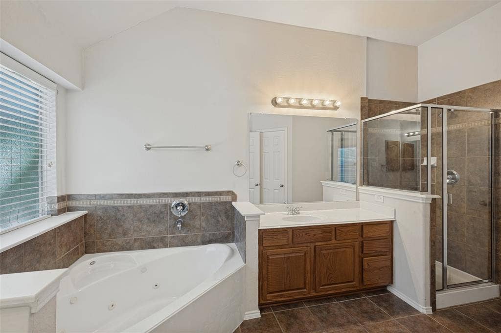 1021 Cherry Hill Lane, DeSoto, TX 75115 Corner jetted tub with tile surround, wood cabinetry vanity, integrated sink, and glass-enclosed shower with tile wall finish