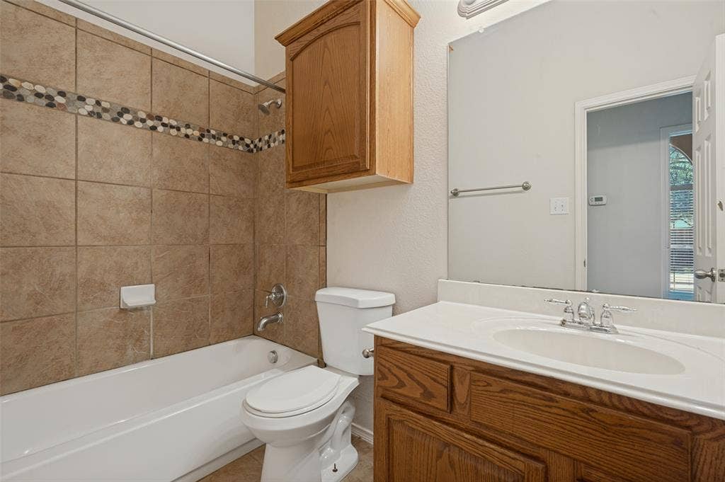 1021 Cherry Hill Lane, DeSoto, TX 75115 Bathroom featuring a vanity with a white countertop and oval under-mount sink, wood-finish cabinetry, wall-mounted storage cabinet, and a tiled shower surround with a decorative accent border