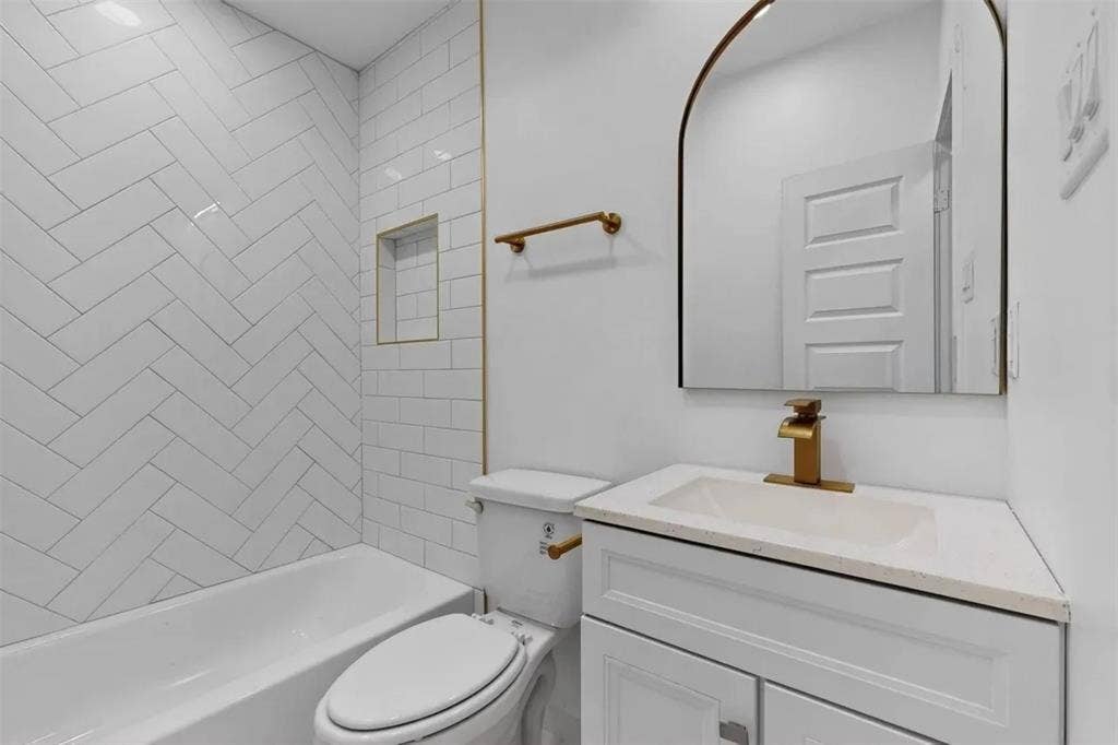 Bathroom featuring a herringbone-patterned tile tub surround, integrated wall niche, arch-top mirror, vanity with stone-composite countertop, and brushed-gold fixtures