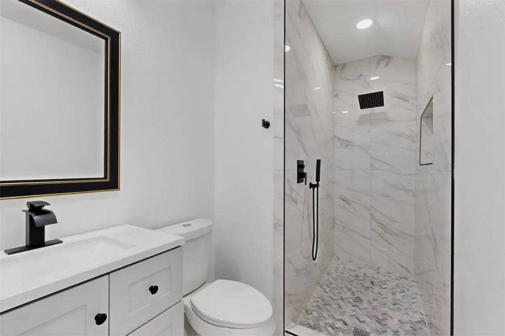 Bathroom featuring a vanity with a white countertop and matte black fixtures, a wall-mounted mirror, and a walk-in shower with stone-patterned wall tile, a herringbone floor inlay, and a built-in recessed shelf