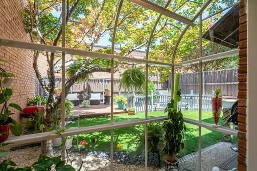 Enclosed sunroom featuring an arched metal-frame glass structure, exposed brick exterior wall, landscaped yard with lawn, raised wooden deck, and privacy fencing