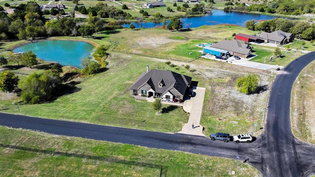 2828 Luke Drive, Farmersville, TX 75442 Aerial perspective of a multi-gabled residence featuring a paved driveway, exterior stone detailing, a pitched shingled roof, and an expansive lot with water features
