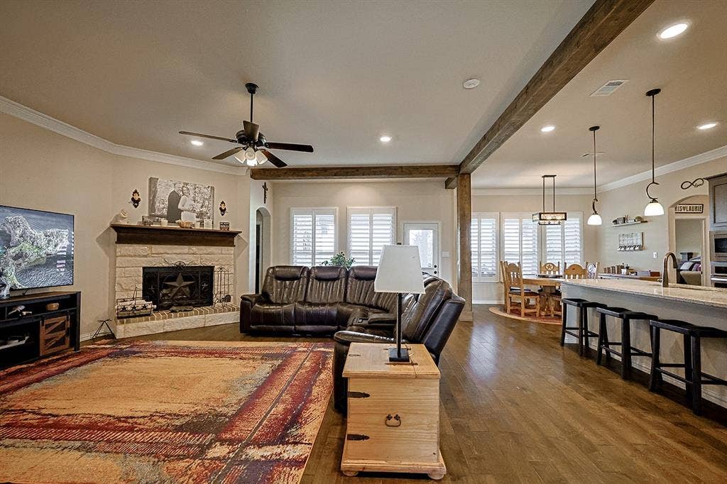 2828 Luke Drive, Farmersville, TX 75442 Open-concept interior featuring wood-finish flooring, exposed ceiling beams, a floor-to-ceiling stone fireplace, plantation shutters, and a kitchen island with pendant lighting