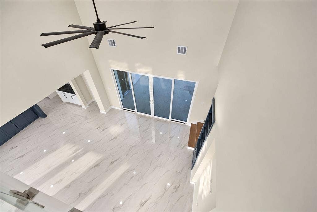 High-ceiling living area featuring large-format stone-look tile flooring, multi-panel sliding glass doors, a modern multi-blade ceiling fan, and staircase with dark metal railing
