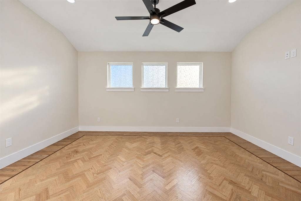 Herringbone wood-finish flooring with a decorative border, vaulted ceiling, three square windows with textured glass, and a matte black ceiling fan with integrated lighting