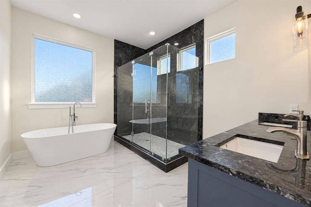 Freestanding white soaking tub with polished chrome floor-mounted faucet, walk-in glass-enclosed shower featuring dark stone-finish surround, dual-sink vanity with dark stone-finish countertop, and large-format tile flooring