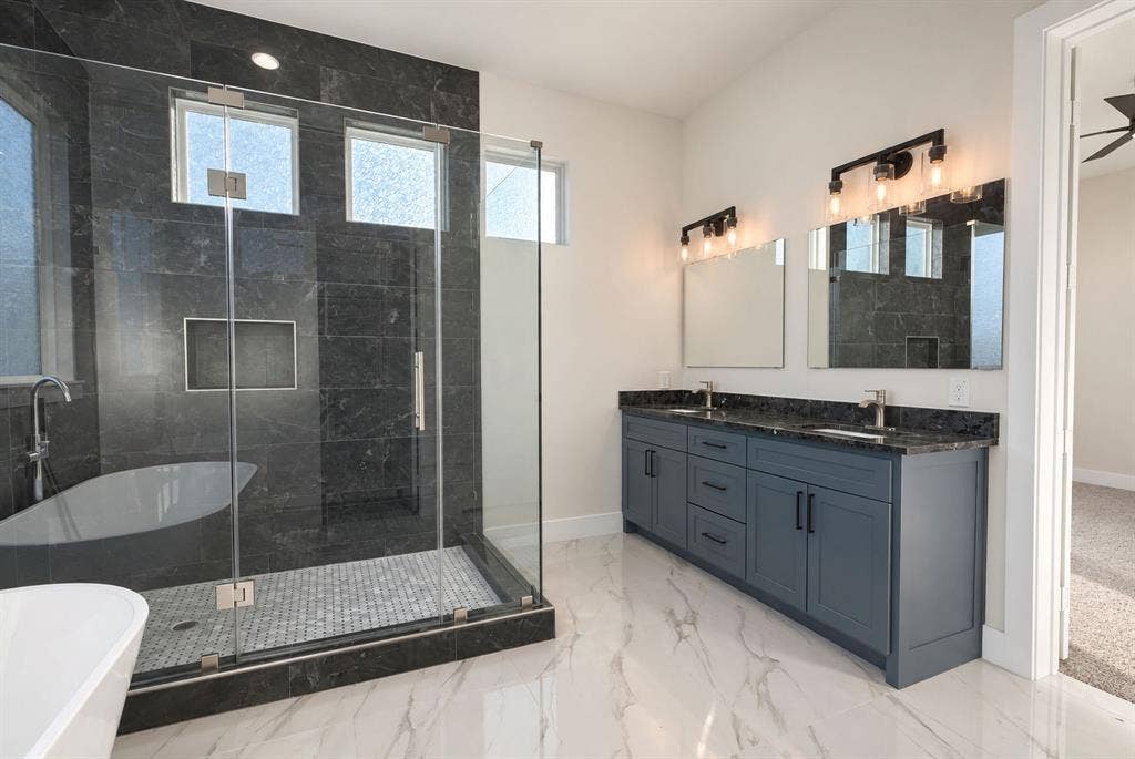 Dual vanity featuring dark cabinetry and a stone-finish countertop, walk-in shower with dark wall tile and integrated shelving, freestanding soaking tub, and polished stone-finish flooring