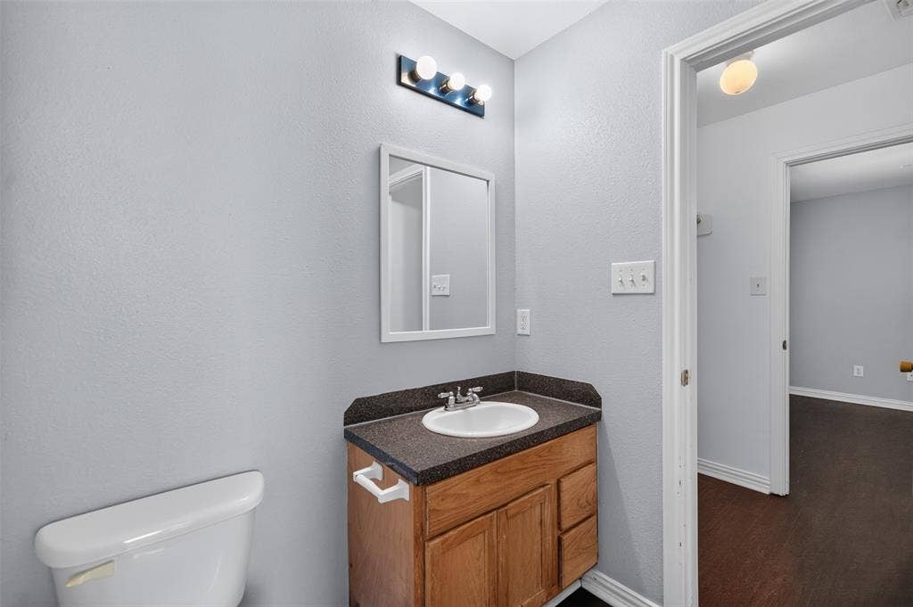1708 Timber View Drive, Tyler, TX 75703 Wood-finish vanity with dark composite countertop, integrated oval basin, white-framed wall mirror, and three-bulb vanity light fixture