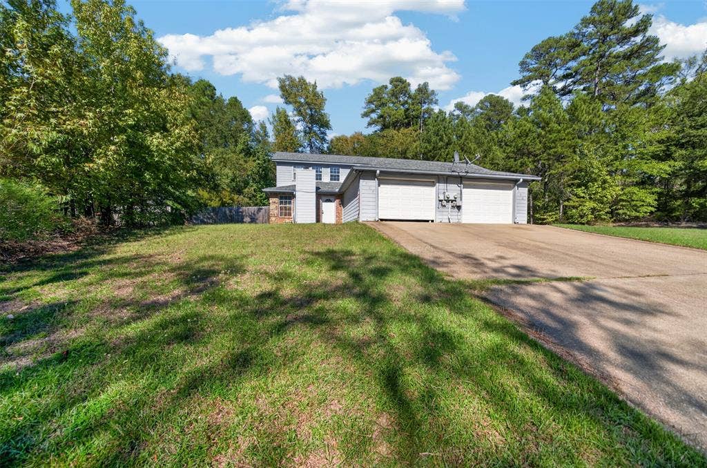 1708 Timber View Drive, Tyler, TX 75703 Two-story residence featuring light-toned horizontal siding, a concrete driveway, a dual-bay garage, and an exterior chimney on a wooded lot