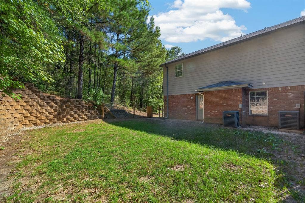 1708 Timber View Drive, Tyler, TX 75703 Rear exterior featuring a brick and horizontal siding facade, tiered stone retaining wall, and wooded perimeter