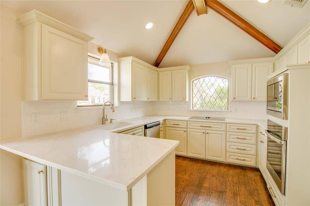 Kitchen featuring shaker-style cabinetry, vaulted ceiling with wood-finish structural beams, quartz countertops, leaded-glass window, and stainless steel built-in double oven