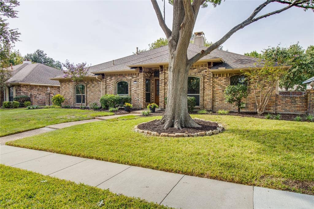 Brick exterior facade featuring an arched entryway, stone-bordered landscaping, prominent mature tree, and a concrete walkway
