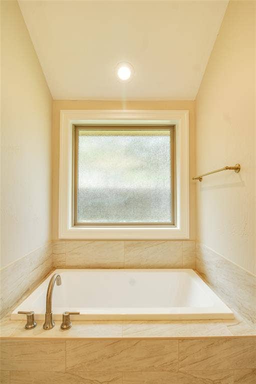 Built-in soaking tub with a metal faucet, surrounded by tile-finish walls and decking, featuring a single-pane window with textured glass and a wall-mounted metal towel rack