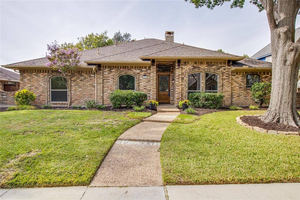 Multi-gabled brick exterior featuring arched windows, a covered entryway with a wood-panel door, a concrete walkway, and a landscaped front lawn with mature trees