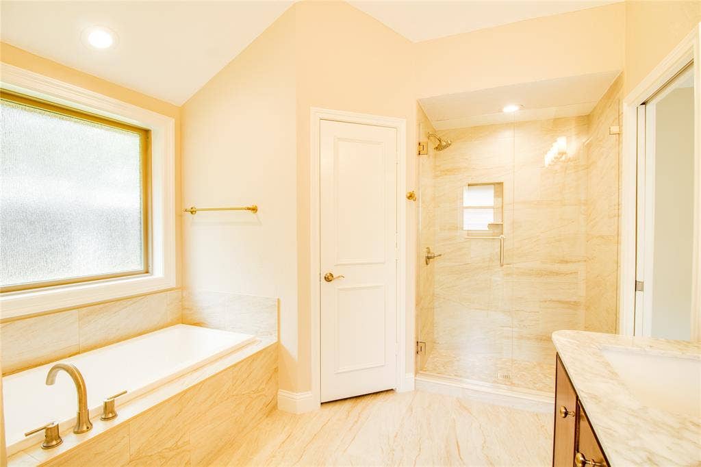 Bathroom featuring a soaking tub, glass-enclosed shower with tiled surround, stone-top vanity, and brass-tone fixtures