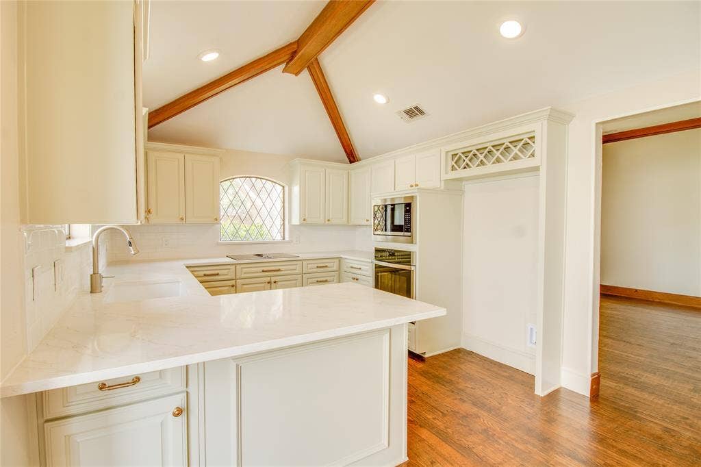Kitchen featuring a vaulted ceiling with exposed wood beams, white cabinetry with built-in wine storage, stone countertops, stainless steel wall-mounted oven and microwave, and wood-finish flooring