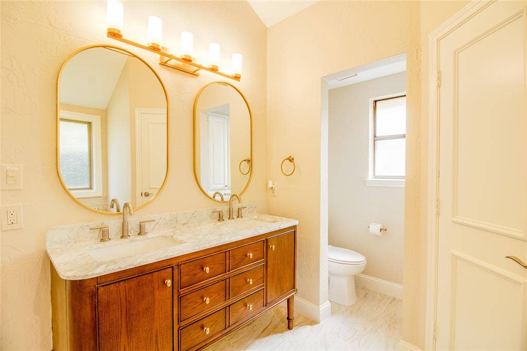 Double vanity with stone countertop, undermount sinks, wood cabinetry, oval wall-mounted mirrors, and four-light gold-toned fixture