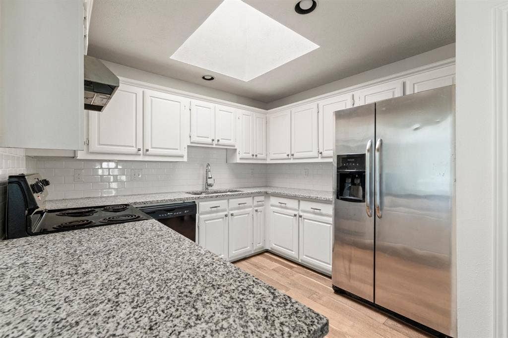 8406 Lullwater Drive, Dallas, TX 75218 Kitchen featuring white shaker-style cabinetry, stone-finish countertops, subway tile backsplash, stainless steel appliances, and a ceiling skylight