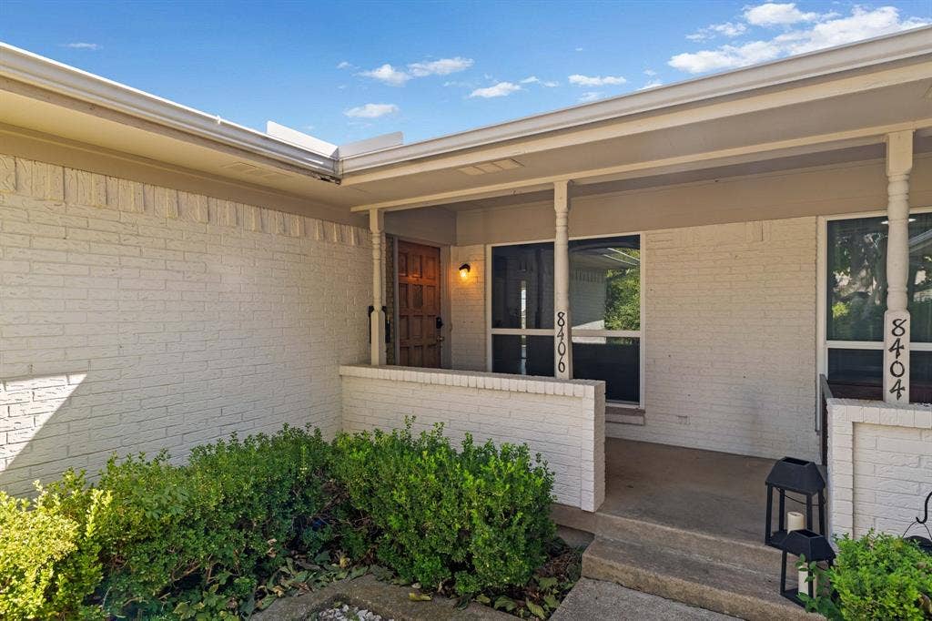8406 Lullwater Drive, Dallas, TX 75218 Painted brick exterior featuring a covered front porch, recessed entryway, custom wood-panel front door, and manicured shrubs