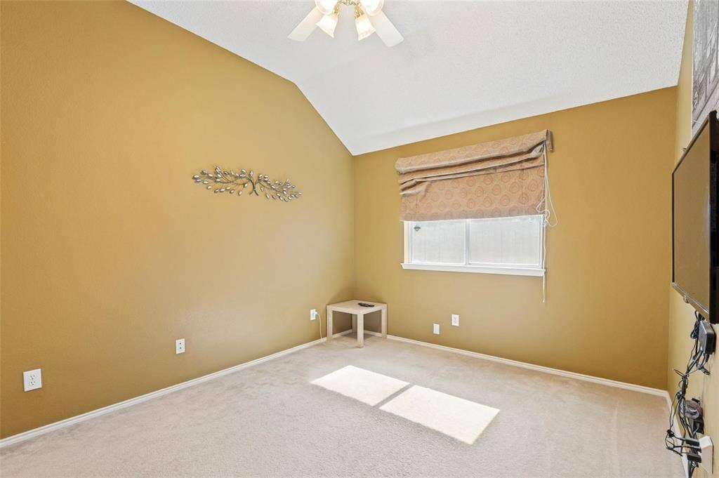 Vaulted ceilings featuring a central overhead fan and light fixture, carpeted flooring, and a window with a fabric roman shade