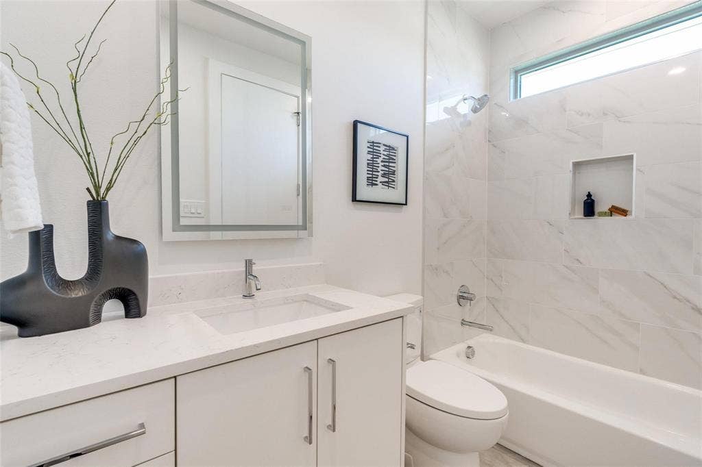 Bathroom featuring a white vanity with a light-toned countertop, integrated rectangular sink, wall-mounted backlit mirror, and a tub-shower combination with floor-to-ceiling stone-look tile, built-in shelving, and transom window