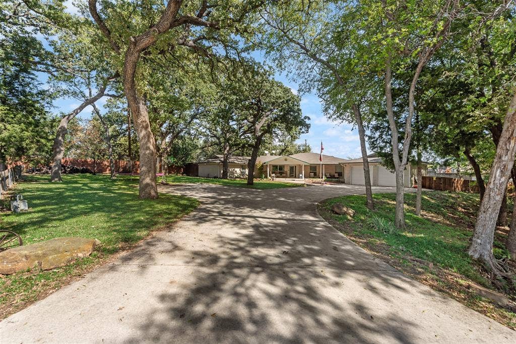 Single-story exterior featuring a concrete driveway, two-car garage, established mature trees, and natural stone landscaping elements