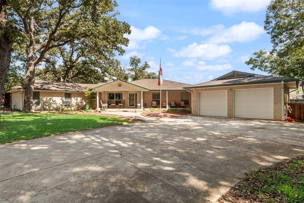 Brick exterior residence featuring a covered front porch, expansive concrete driveway, two-car garage, and mature trees