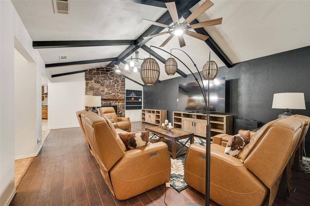 Vaulted ceiling with exposed decorative beams, stone-faced fireplace, and wood-finish flooring