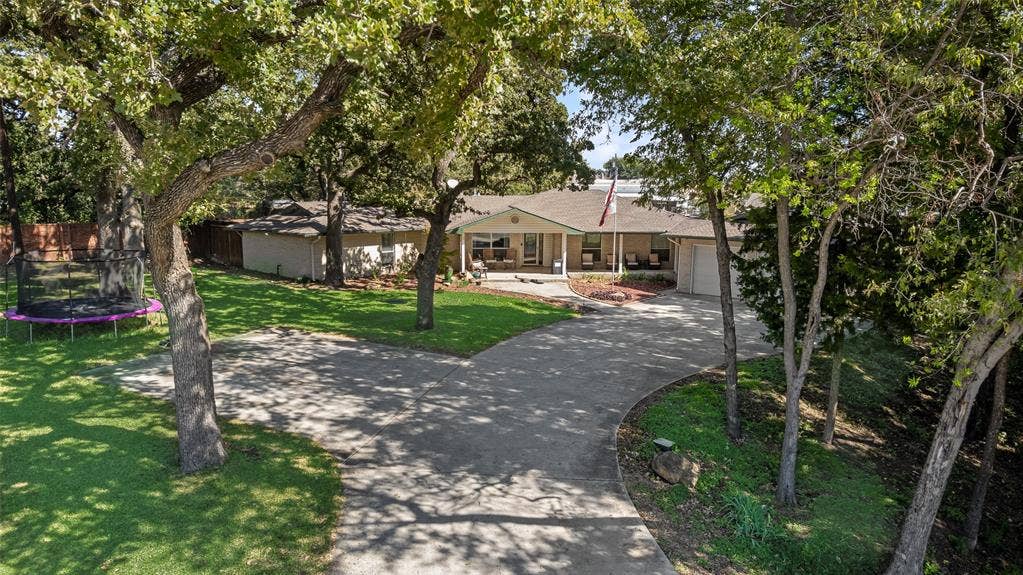 Single-story exterior featuring a brick facade, covered front porch, attached garage, curved concrete driveway, and established mature trees