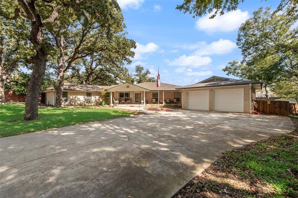 Single-story brick exterior featuring a covered front porch, an attached two-car garage, expansive concrete driveway, and mature trees on a landscaped lot