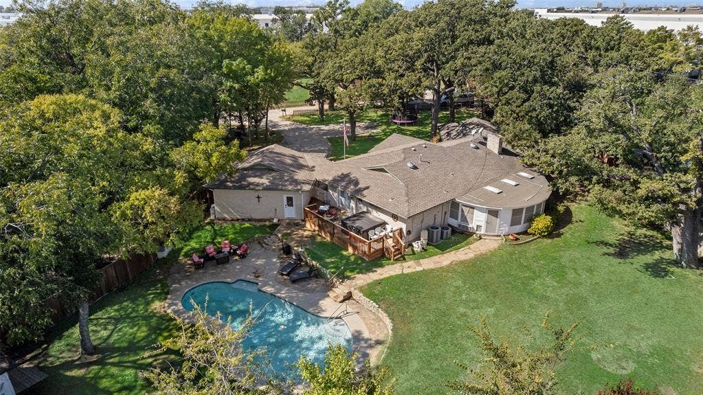 Aerial perspective of a sprawling property featuring a swimming pool, stone-surfaced patio, elevated wood decking, and mature trees across a grass landscape