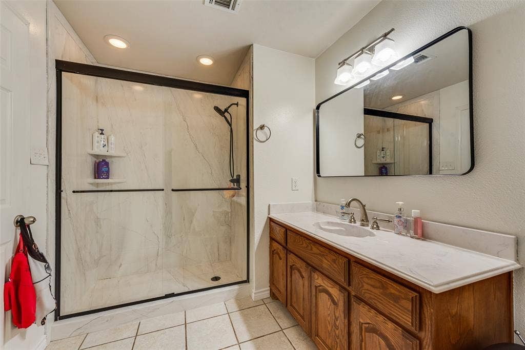 Walk-in shower with floor-to-ceiling stone-pattern tile surround and matte black hardware, vanity featuring a solid surface countertop, wood cabinetry, and a large rectangular mirror with a matte black frame