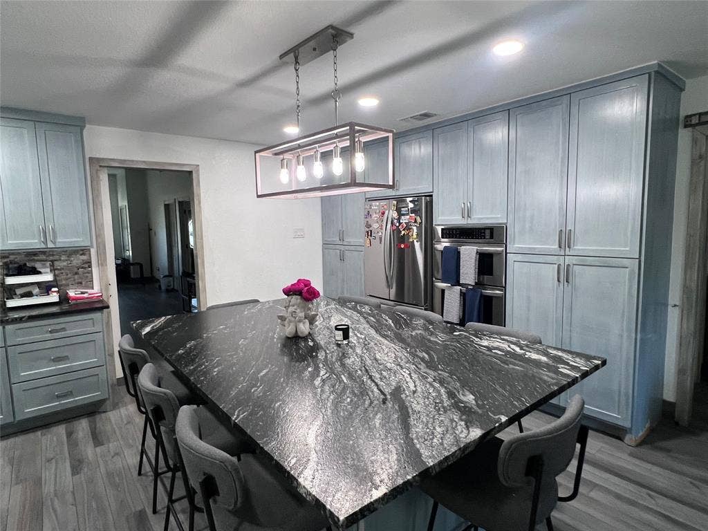201 Darien Drive, Mesquite, TX 75149 Kitchen featuring custom light blue cabinetry, a large stone-topped island, a linear multi-bulb pendant light fixture, stainless steel double wall ovens, and wood-finish flooring