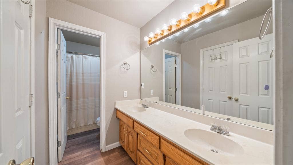 8935 Bluebonnet Drive, Scurry, TX 75158 Bathroom vanity featuring a light-toned countertop with integrated dual sinks, wooden cabinetry, a full-width wall mirror, and a linear light fixture