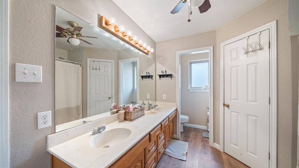 8935 Bluebonnet Drive, Scurry, TX 75158 Double vanity featuring a light-toned countertop with integrated dual sinks, wood-finish cabinetry, full-width wall mirror, and wood-finish flooring