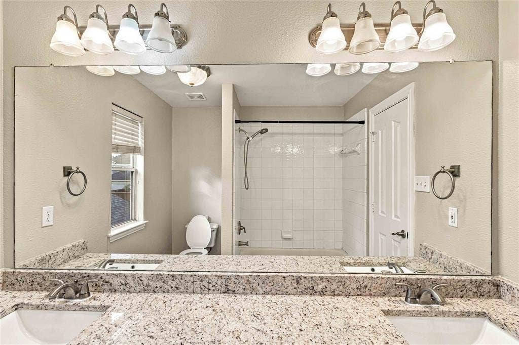 Dual-sink vanity with stone countertop, full-width vanity mirror, dual four-light fixtures, and white tile shower surround with a handheld showerhead