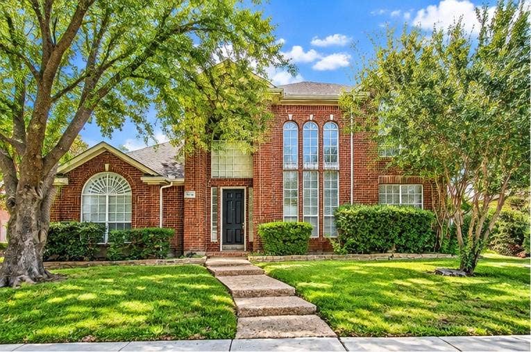 Two-story brick exterior featuring a prominent arched window, multi-pane windows, a concrete walkway, and mature landscaping