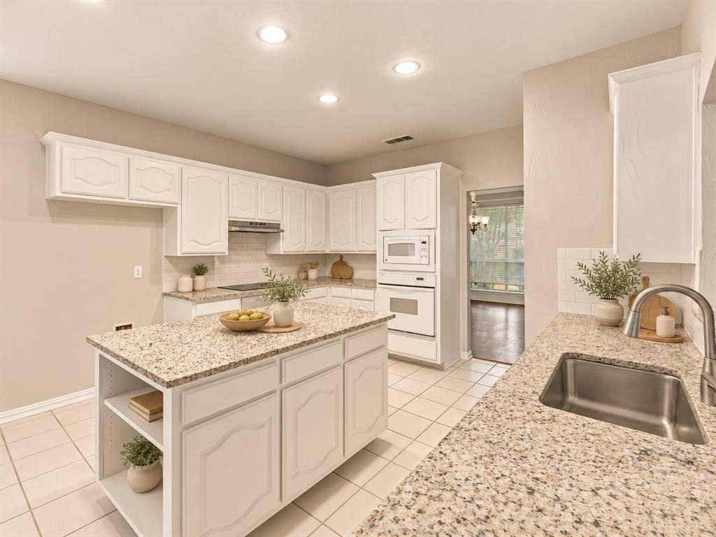 Kitchen featuring white cabinetry, stone-finish countertops, a central island with integrated shelving, built-in wall oven and microwave, and ceramic tile flooring