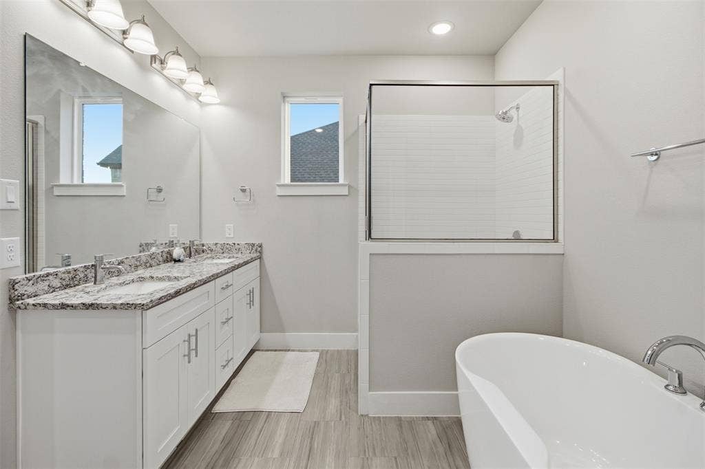 1408 Rockcliff Street, Plano, TX 75093 Double vanity featuring stone countertops and integrated sinks, accompanied by a freestanding soaking tub, a tiled glass-enclosed shower, and wood-finish flooring