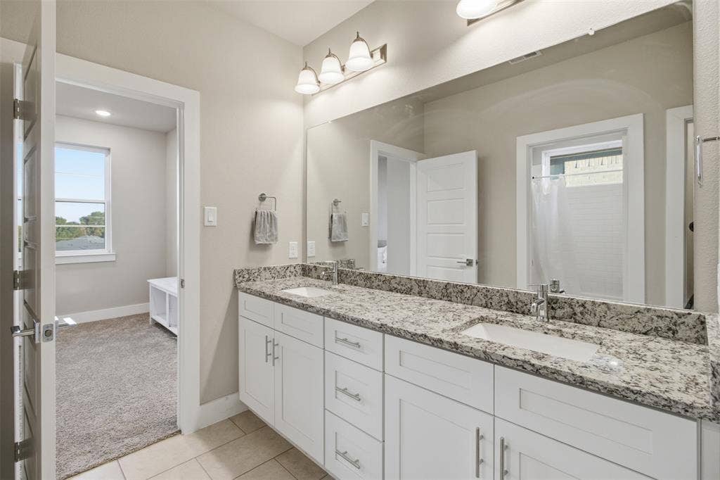 1408 Rockcliff Street, Plano, TX 75093 Dual-sink vanity with stone-finish countertop, white shaker-style cabinetry, large wall-mounted mirror, and three-bulb vanity lighting