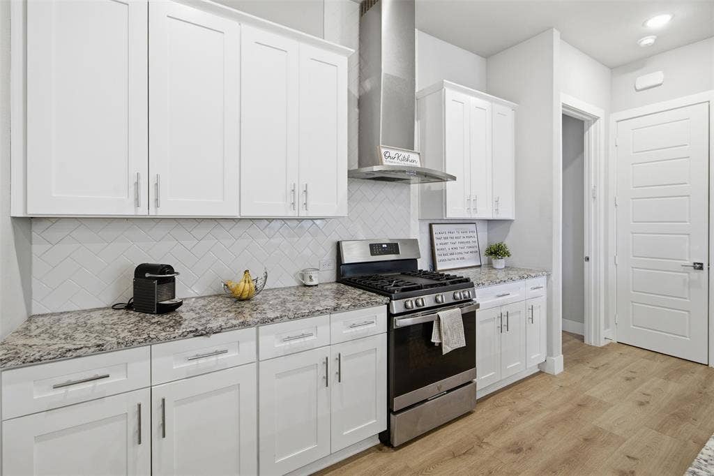 1408 Rockcliff Street, Plano, TX 75093 Kitchen featuring white shaker-style cabinetry, stone-finish countertops, herringbone-pattern tile backsplash, stainless steel range with matching vent hood, and wood-finish flooring