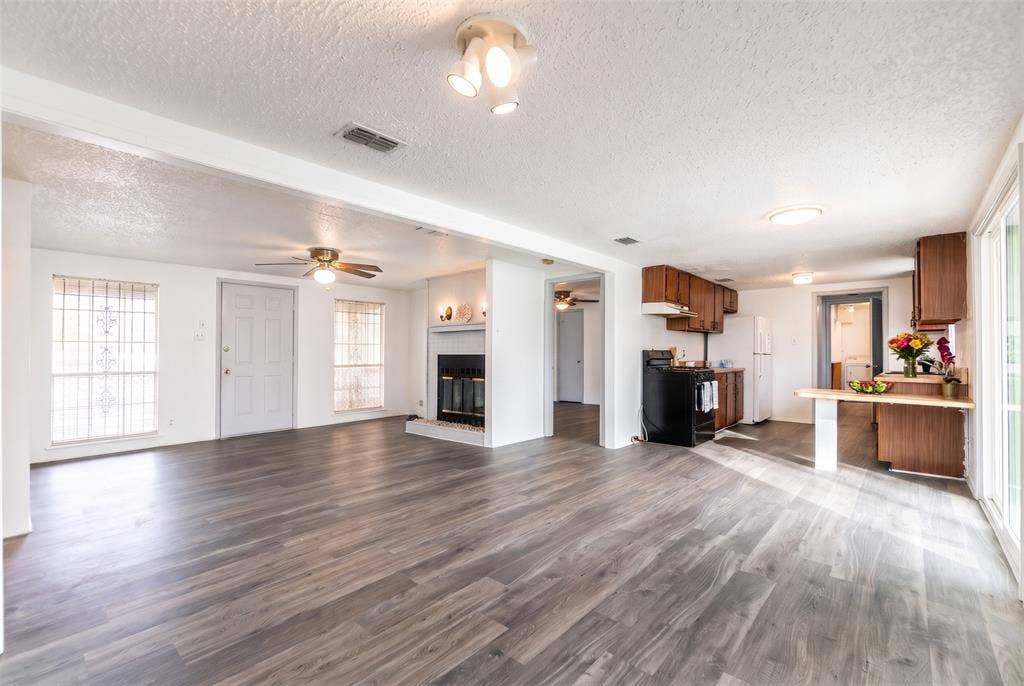 Open-concept interior featuring wood-finish flooring, a central fireplace with a brick surround, a multi-light ceiling fixture, and wood cabinetry