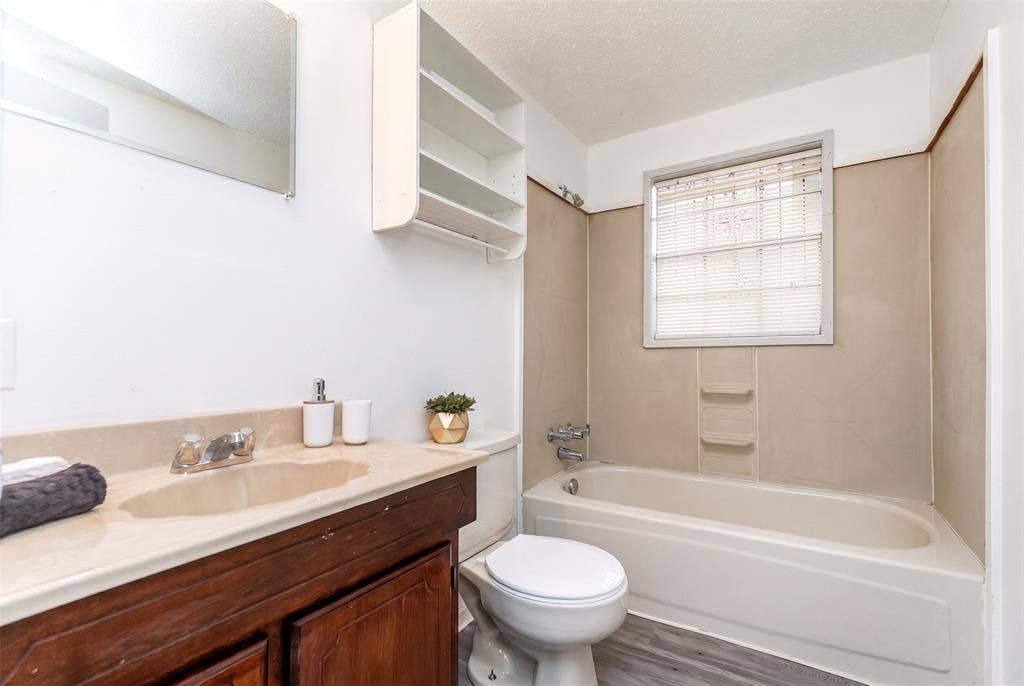 Bathroom featuring a wood-finish vanity with an integrated countertop and sink, wall-mounted storage shelving, a combination bathtub and shower enclosure with molded shelving, and wood-finish flooring